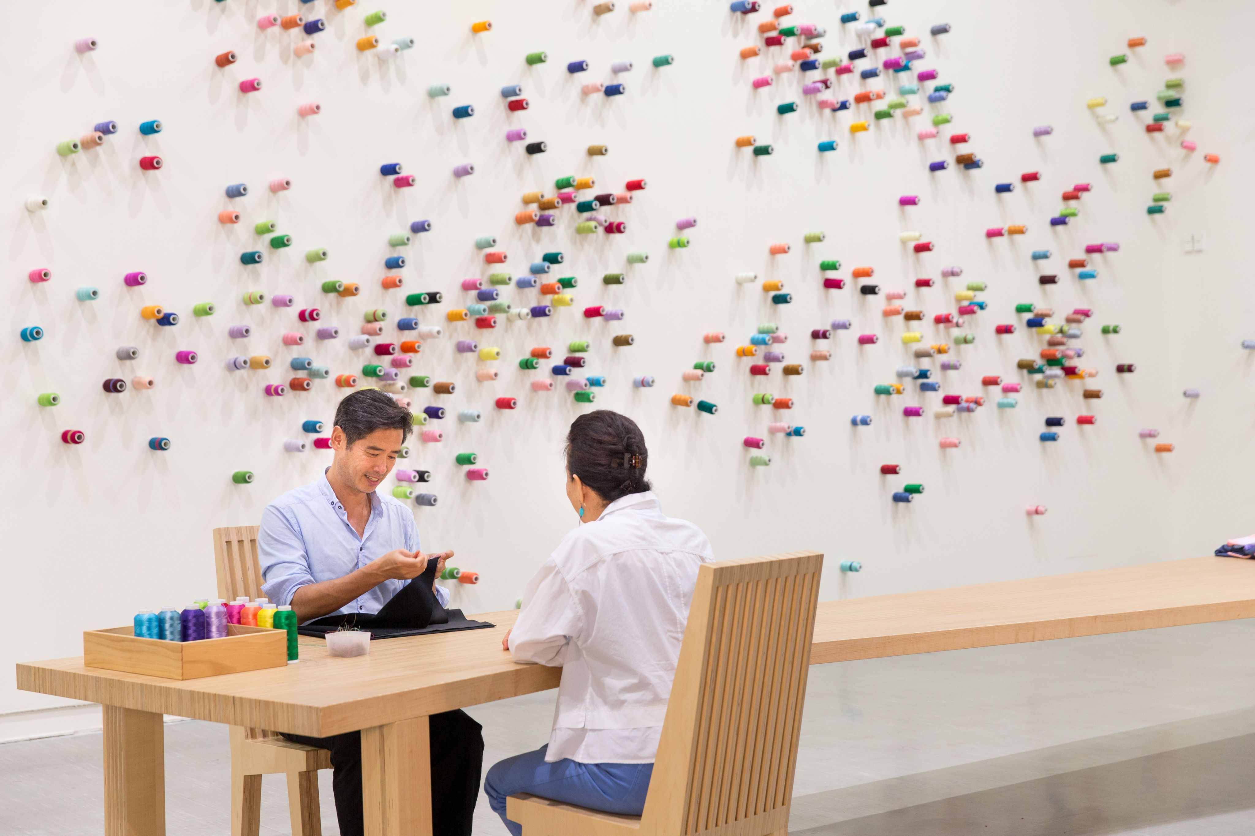 An artist sits at a table with an audience and brightly colored spools of thread on the wall