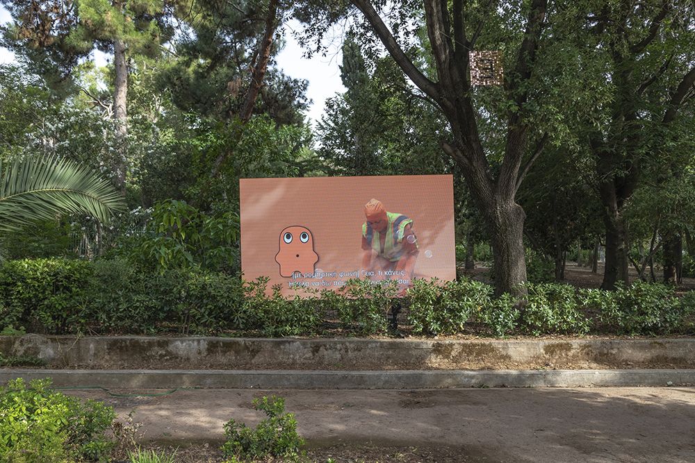 A screen showing a figure in all orange kneeling next to an animated orange blob is set up in a lush, tropical park