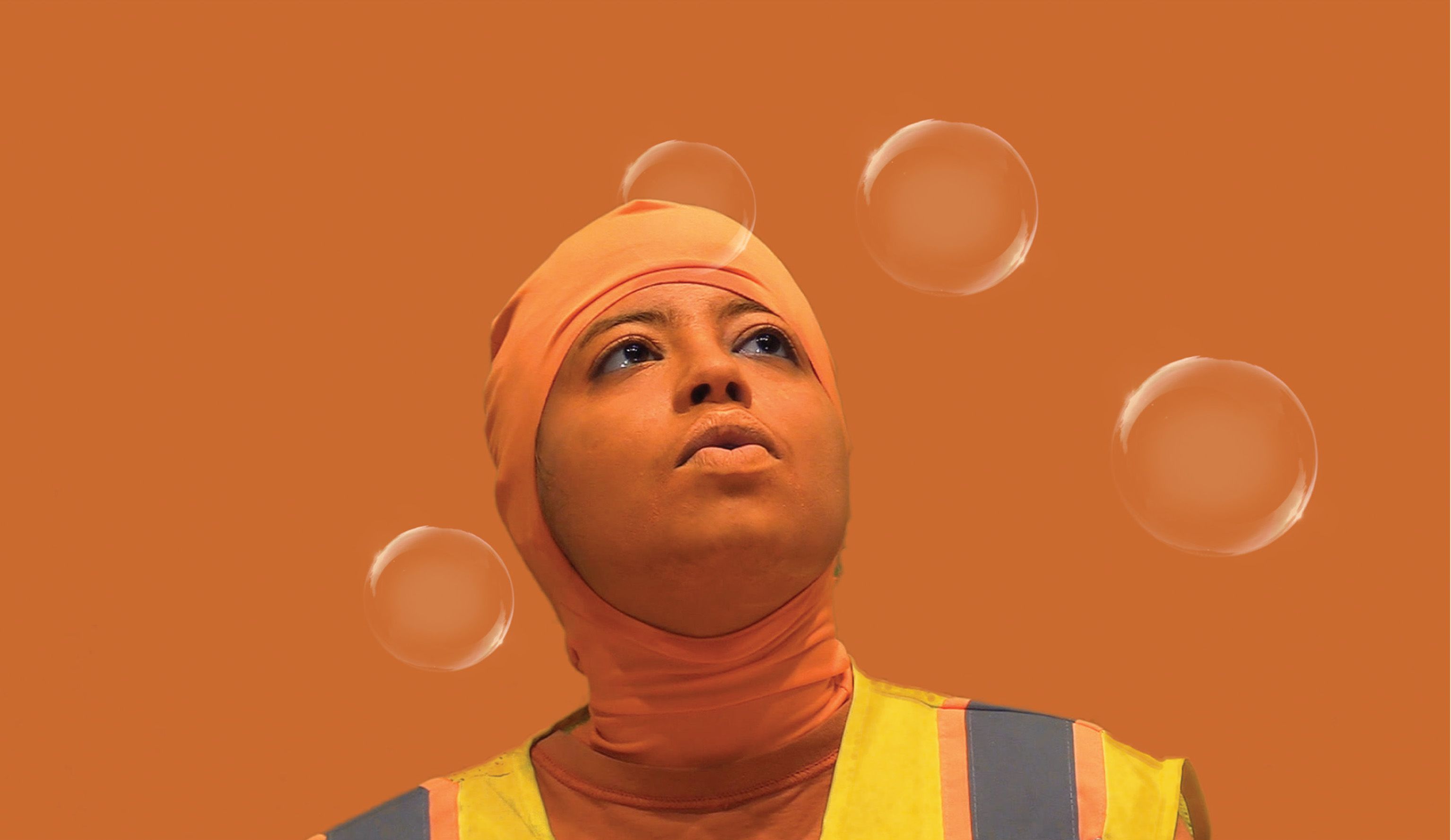 A close-up of a person who is wearing an orange baclava, their skin painted orange, with bubbles surrounding their head, standing in front of an orange background