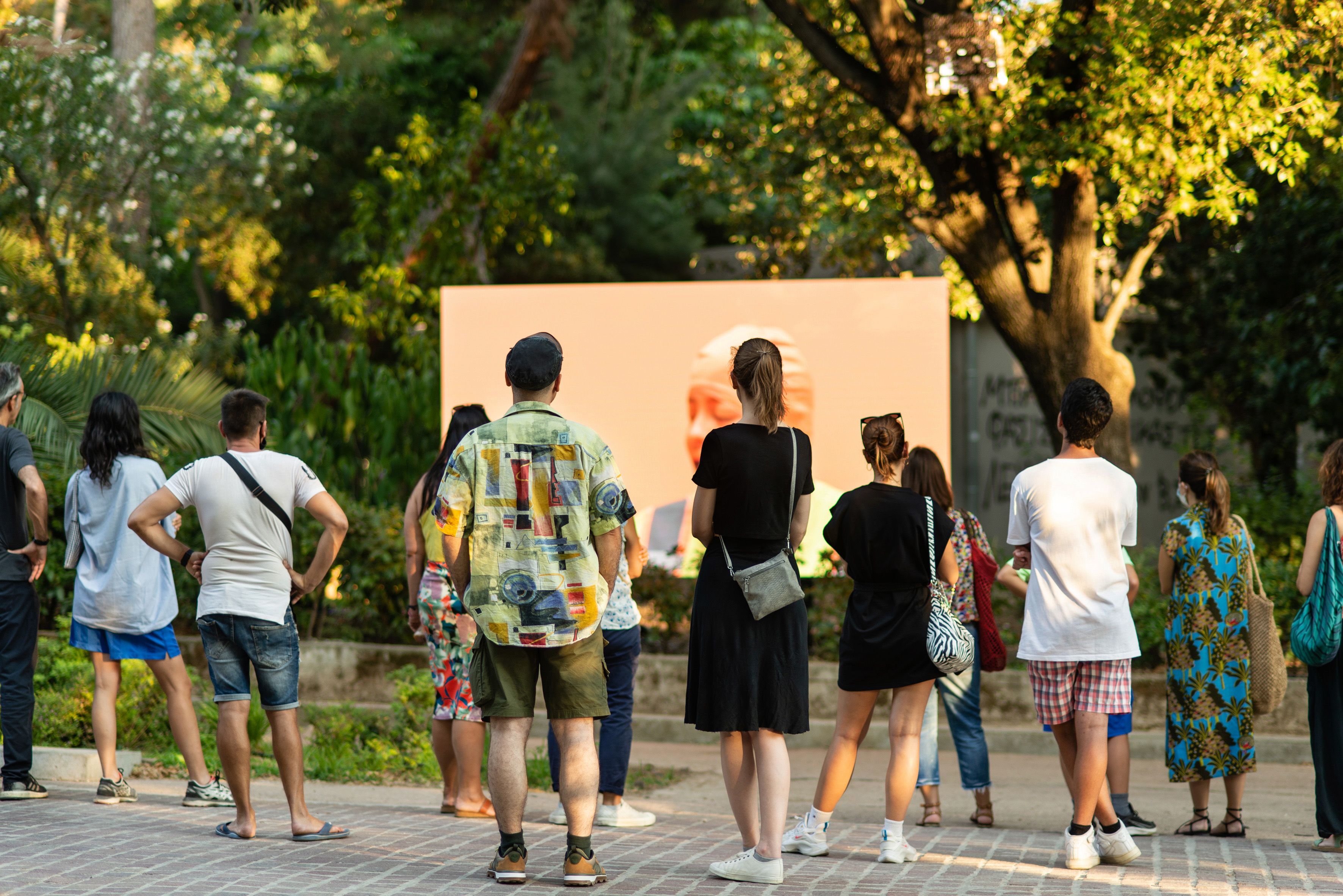 A crowd stands in front of a screen showing an orange backdrop and the artist's head, set up in a lush park