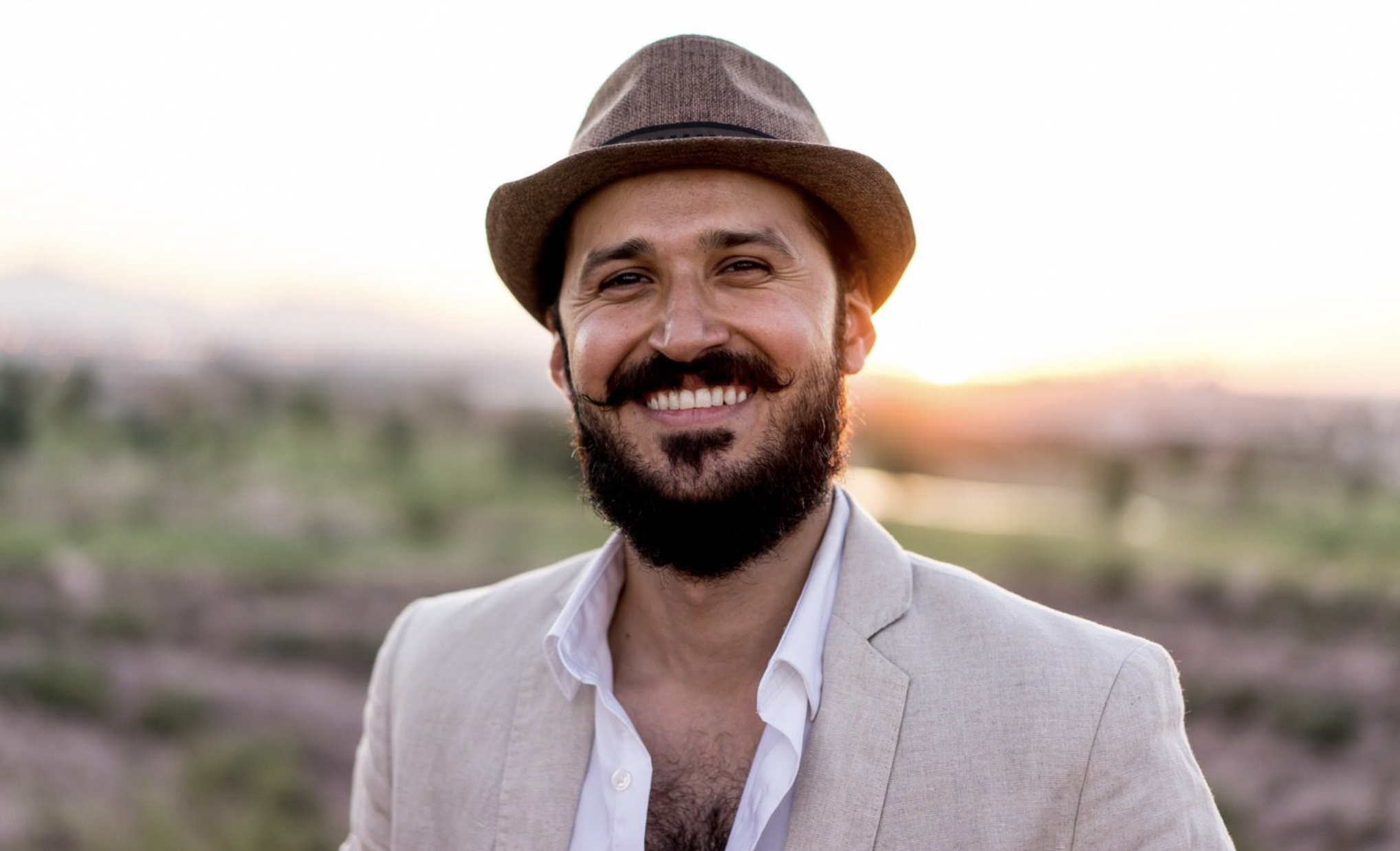 Bassim Al-Shaker wears a brown hat, a tan blazer, an unbottoned white collared shirt, and a smile. He is standing in front of the sun as it rises above the horizon.