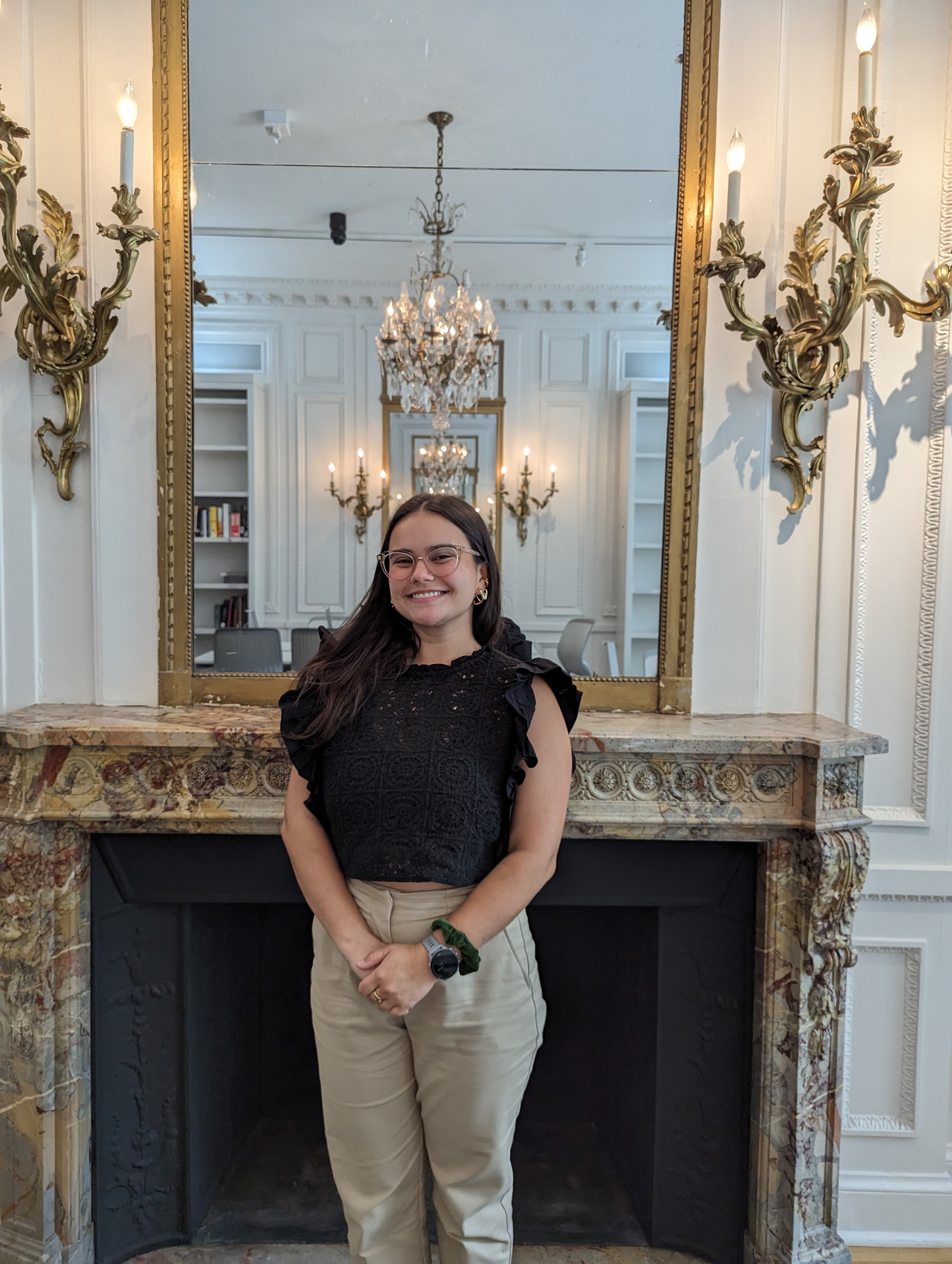 A young woman stands in front of an ornate mirror and fireplace