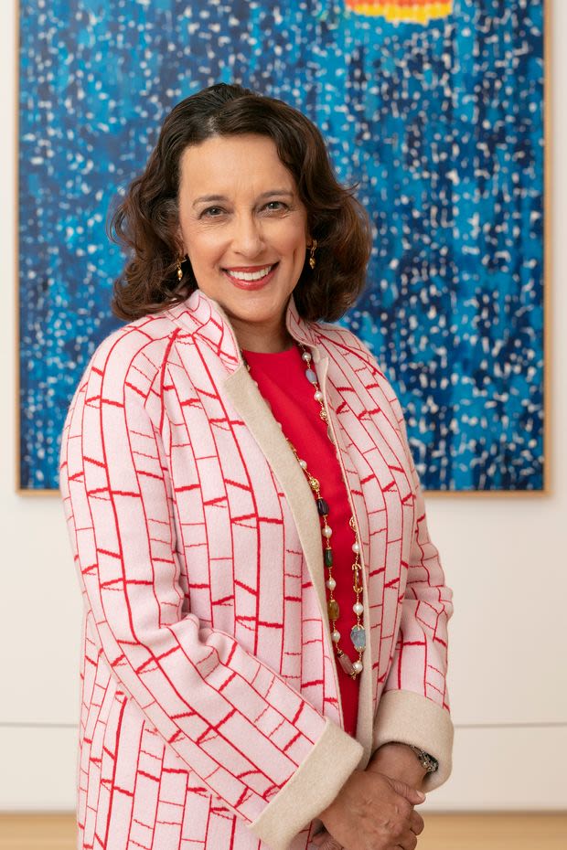 A woman with shoulder-length brown hair, wearing a red-and-white coat, a red top, and round multicolored beads that extend past the length of the photo smiles at the camera in front of a dark blue painting depicting trees on a starry night.