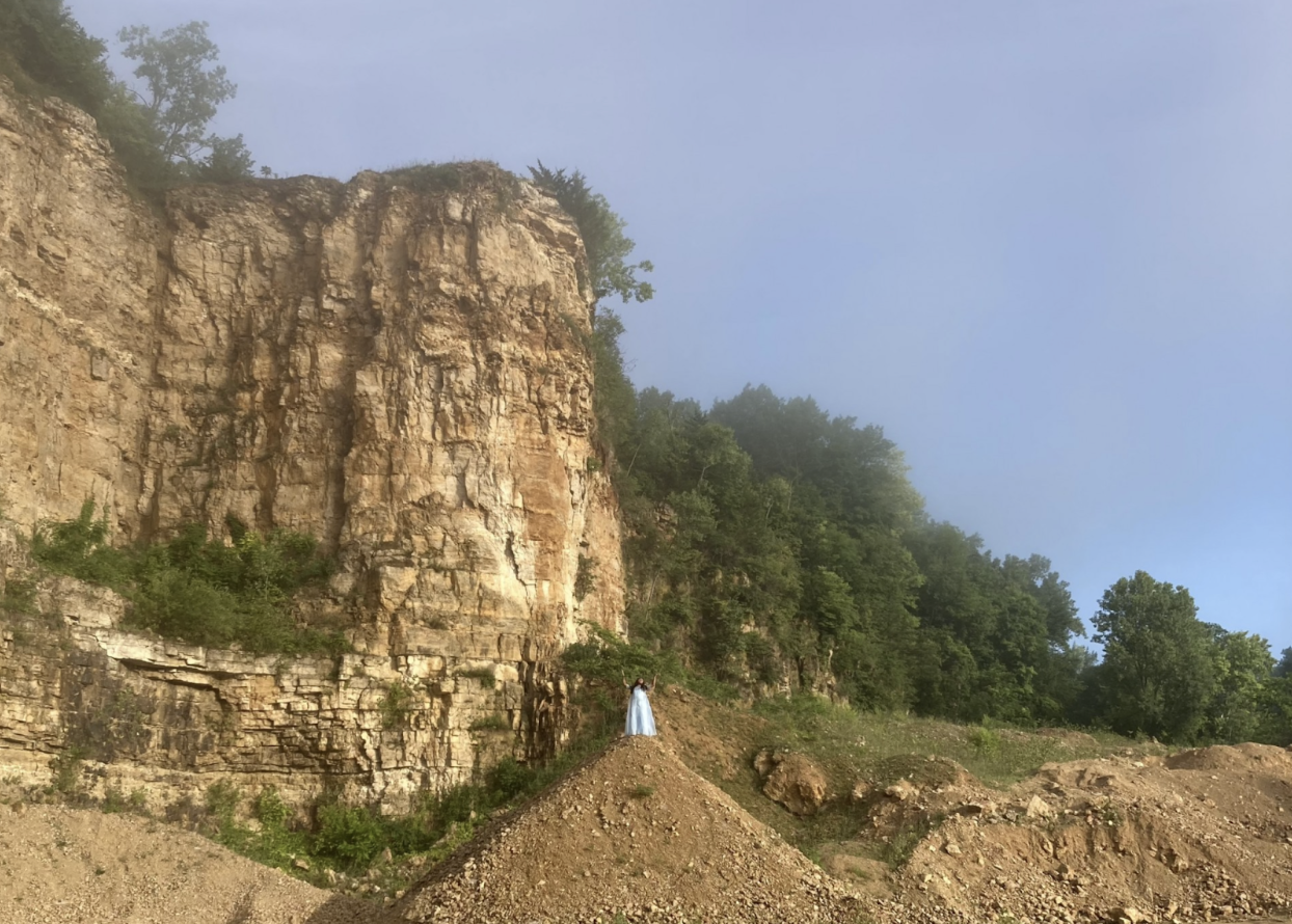 A person wearing a white dress stands on a large mound of dirt. Behind them is a tall rocky cliff and a gathering of large trees.