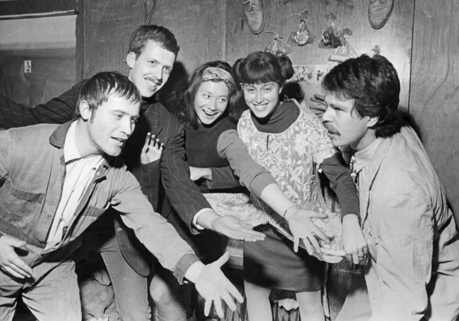 Five group of people (two women and three men) stand close together. Four members of the group reach out their hands to the man standing on the far right of the group with smiles on their faces. The picture is taken in black and white.