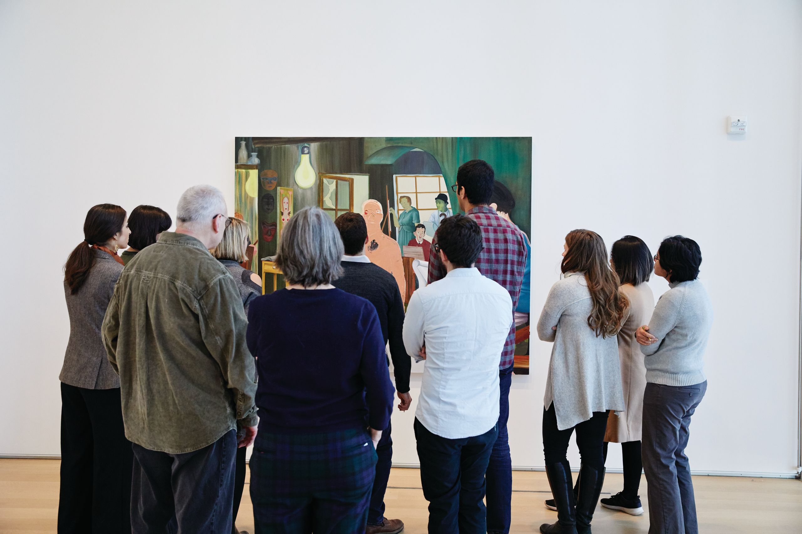 A group of people face away from the camera and ponder a painting hanging on a white wall