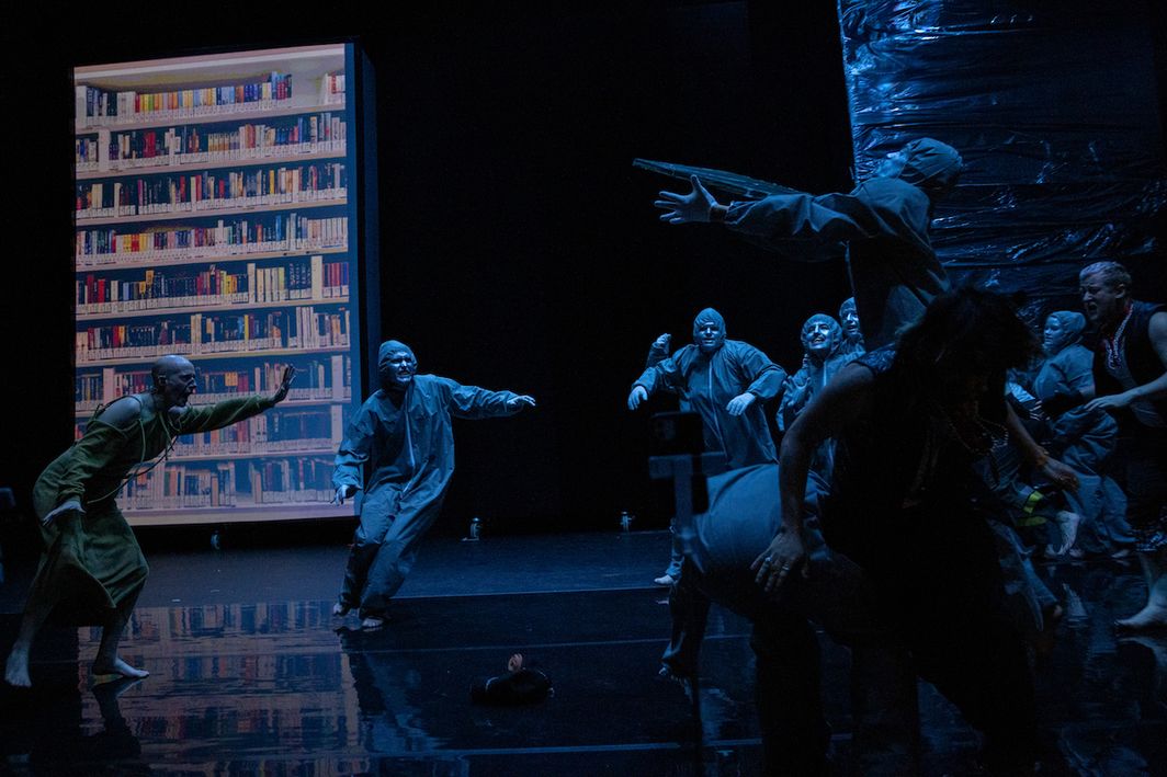 Several figures in hooded jumpsuits on a stage. Many are reaching towards each other. The lighting is blue and there is a large image of a library shelf in the background.