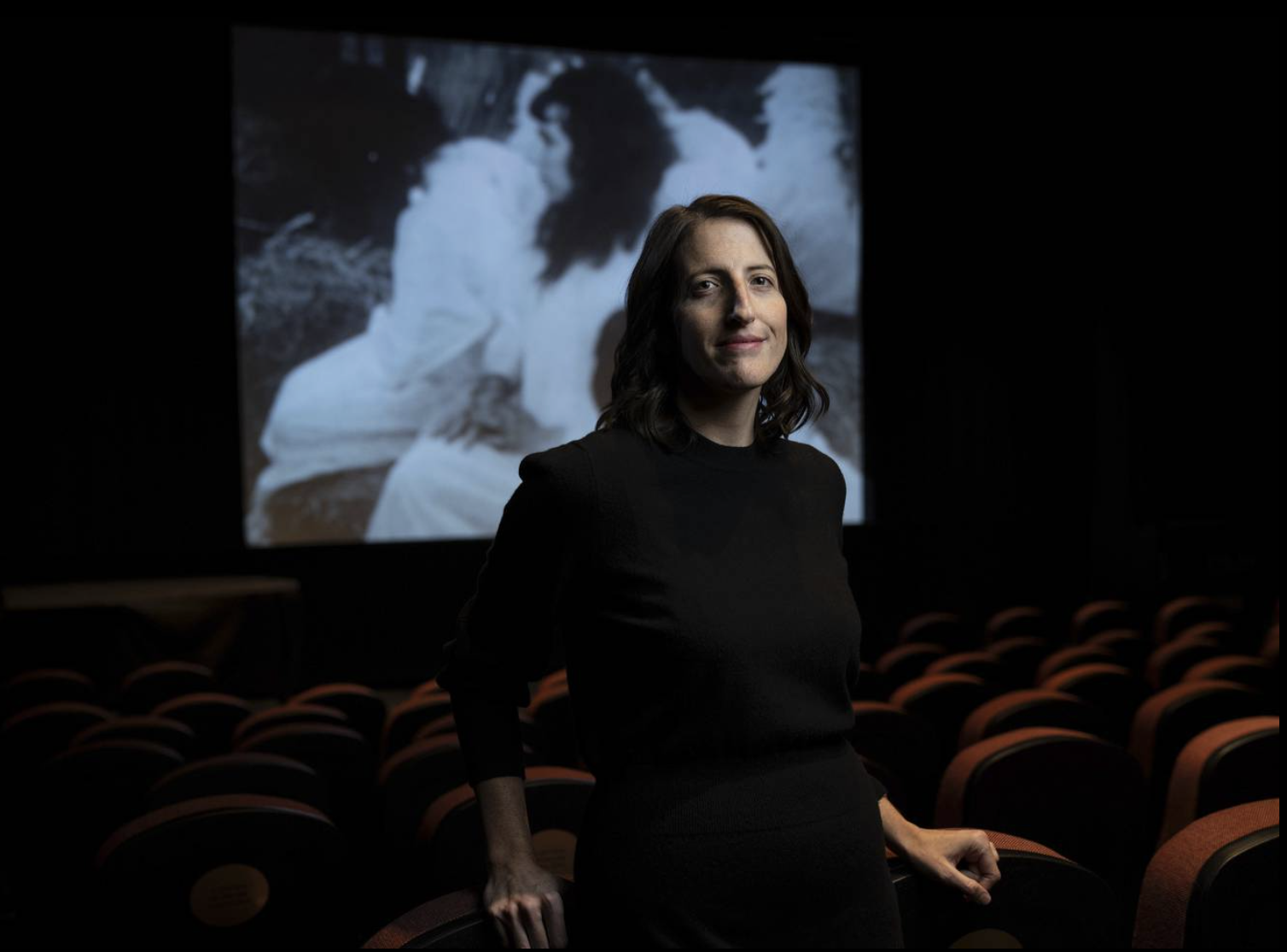 Rebecca Fons stands in a dark movie theater room in the middle of rows of red movie theater chairs. On the screen behind her plays black-and-white film, which shows a still of a woman wearing white clothes.