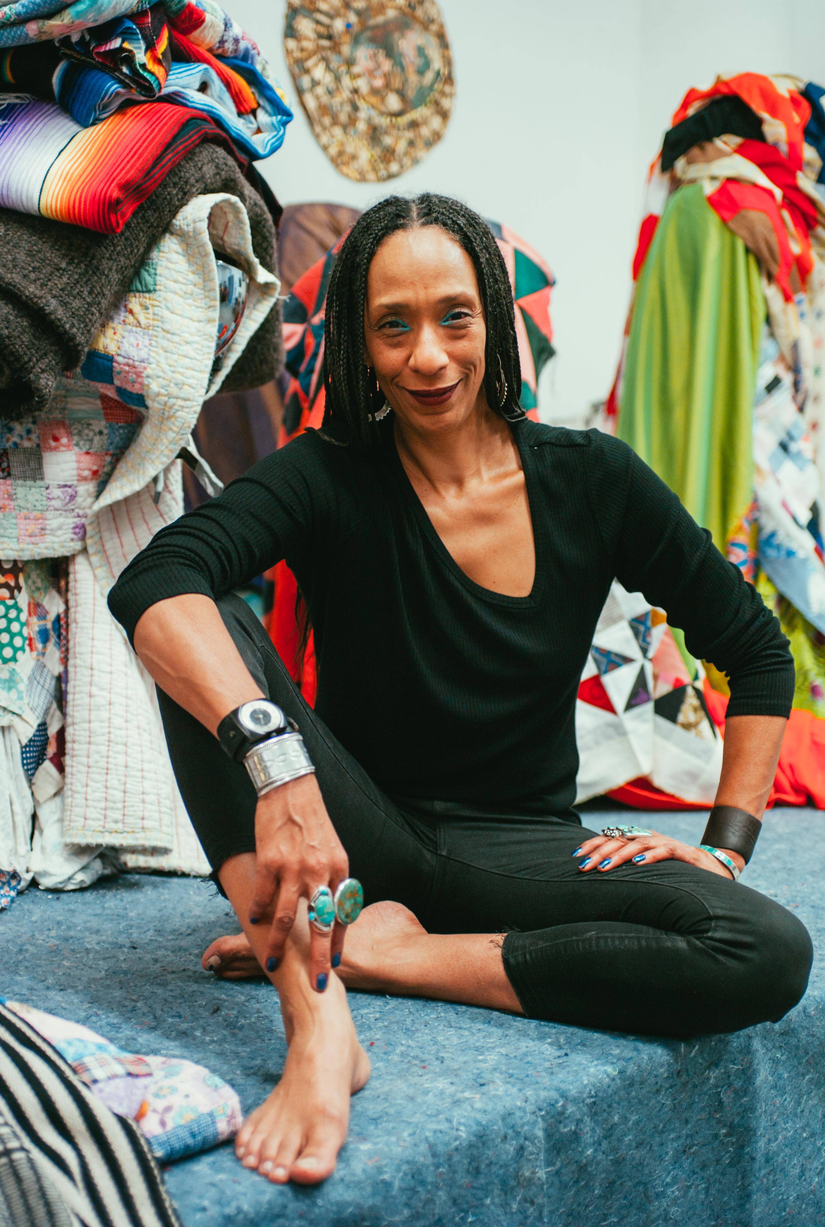 A woman dressed all in black sits on the blue carpet of her studio, surrounded by bright, patterned textiles