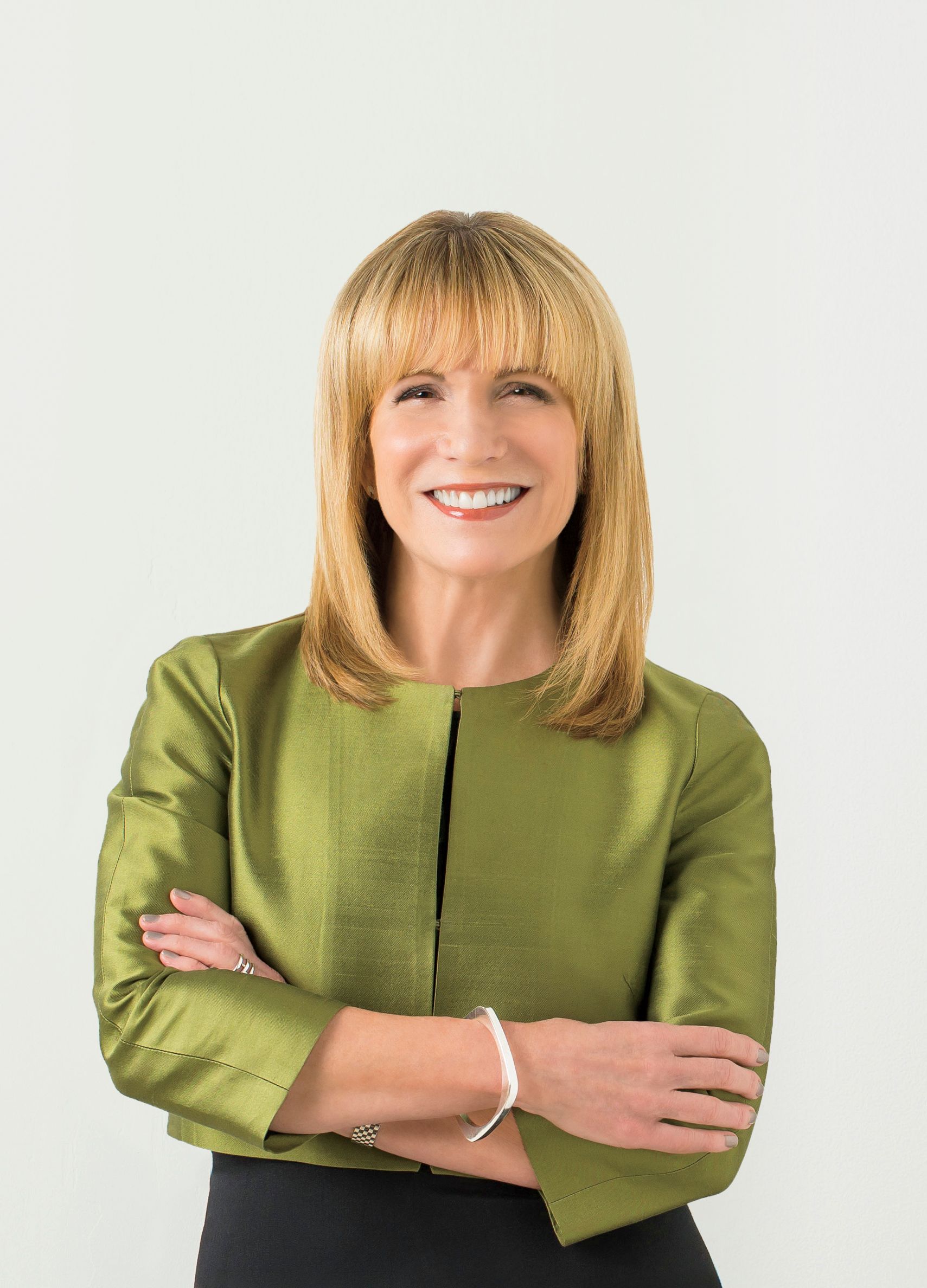 A white woman with shoulder-length blonde hair and straight-across bangs, wearing a chic olive green structured blazer, crosses her arms and smiles into the camera