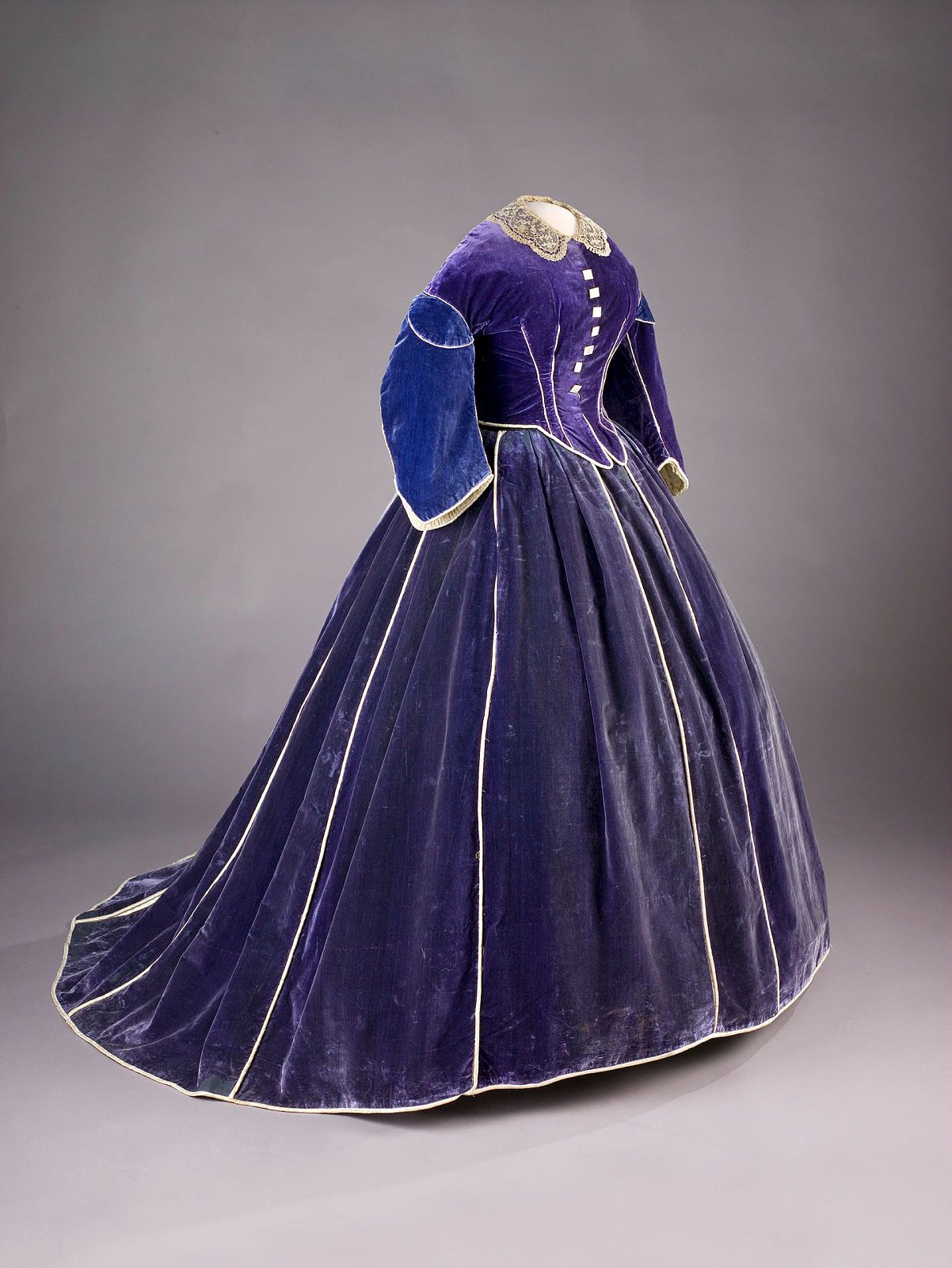 A mannequin dressed in a purple velvet dress stands in front of a gray and white background. The dress has white satin piping, mother of pearl buttons, and an intricate, lace collar.