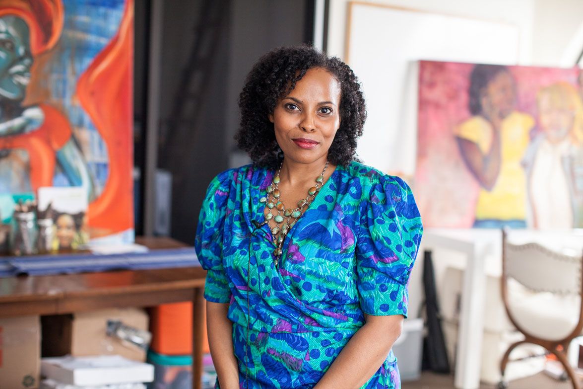 A photograph of artist Scheherazade Tillet. Tillet is wearing a blue, green, and purple dress and is looking at the camera. An artist studio is visible behind her.