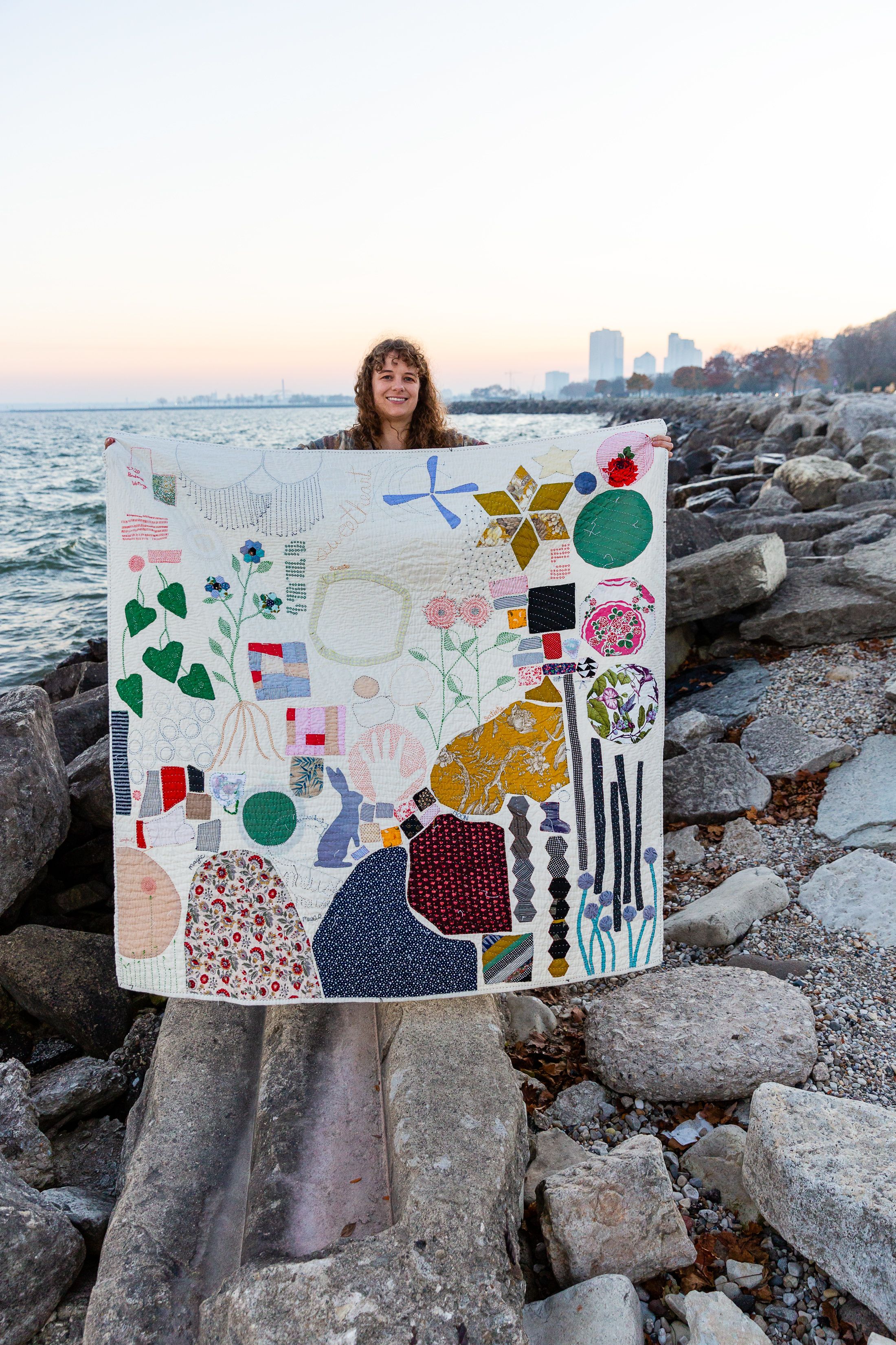 An artist holds a piece of textile art up on a stone pier