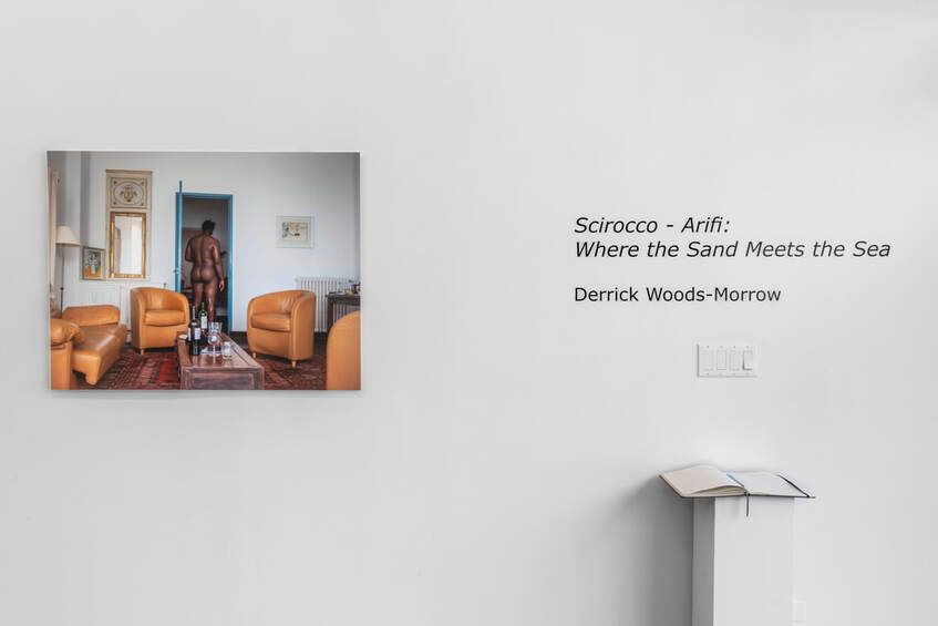 Scirocco - Arifi: Where the Sand Meets the Sea is written on a gallery wall next to a photograph of a nude man
