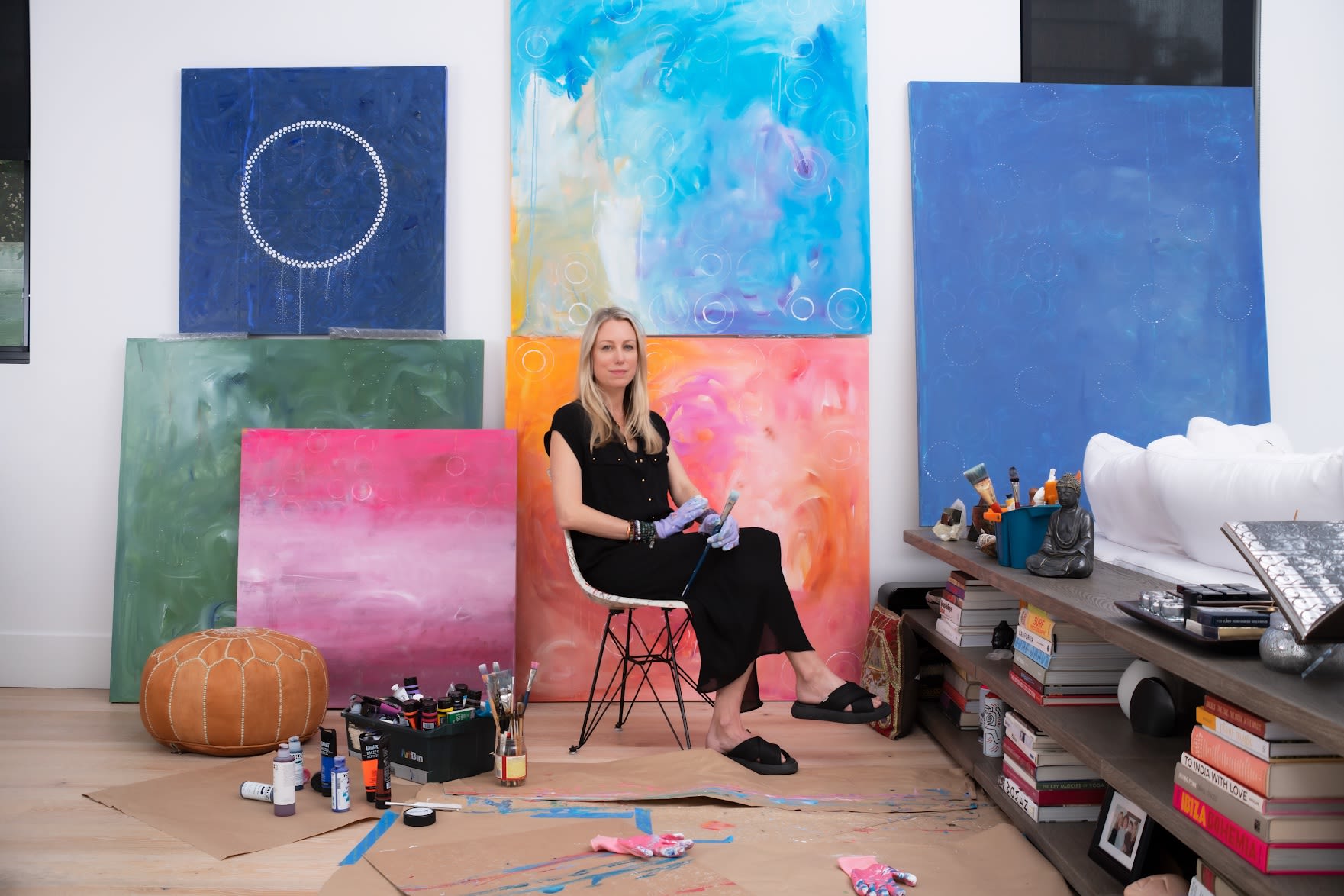 An artist sits in their studio with large colorful paintings against the wall