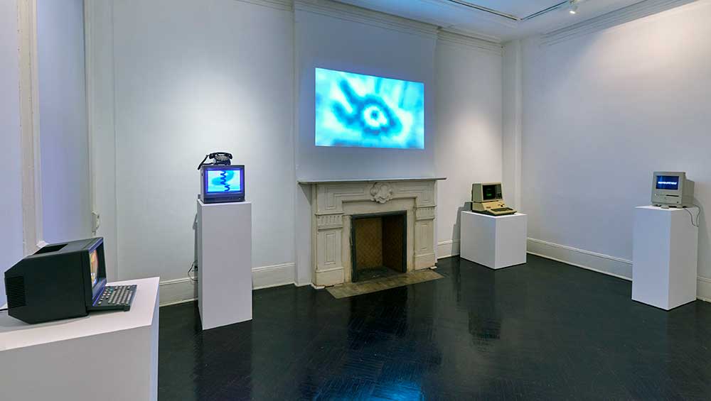 An exhibition view of Eduardo Kac: From Minitel to NFT featuring computer screens on pedestals and a glowing screen of a large, pixelated eye