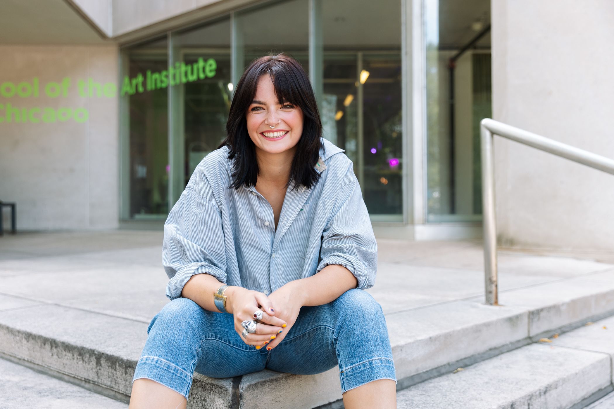 A student in a button-up shirt and jeans sits outside a campus building