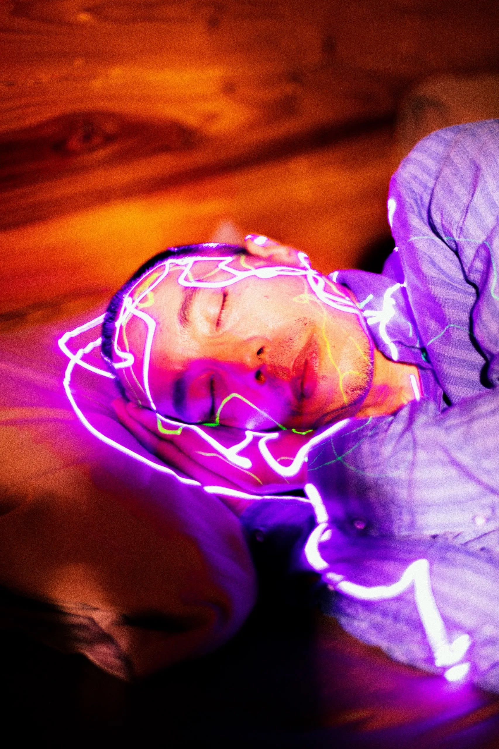Artist Apichatpong Weerasethakul is photographed laying down on a whimsical orange background. He is wearing a purple striped shirt and organic neon purple and green lines surround his head and upper body.