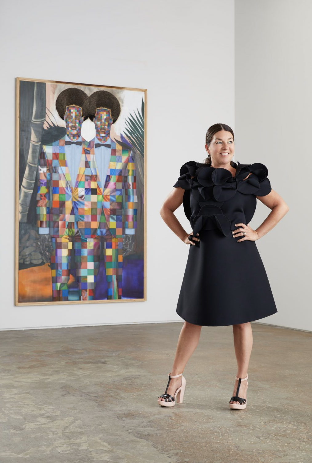A woman wearing a black dress stands in front of a painting of two Black people with colorful outfits and similarly-styled afros with both hands on her hips. She looks off to the side with a smile on her face.