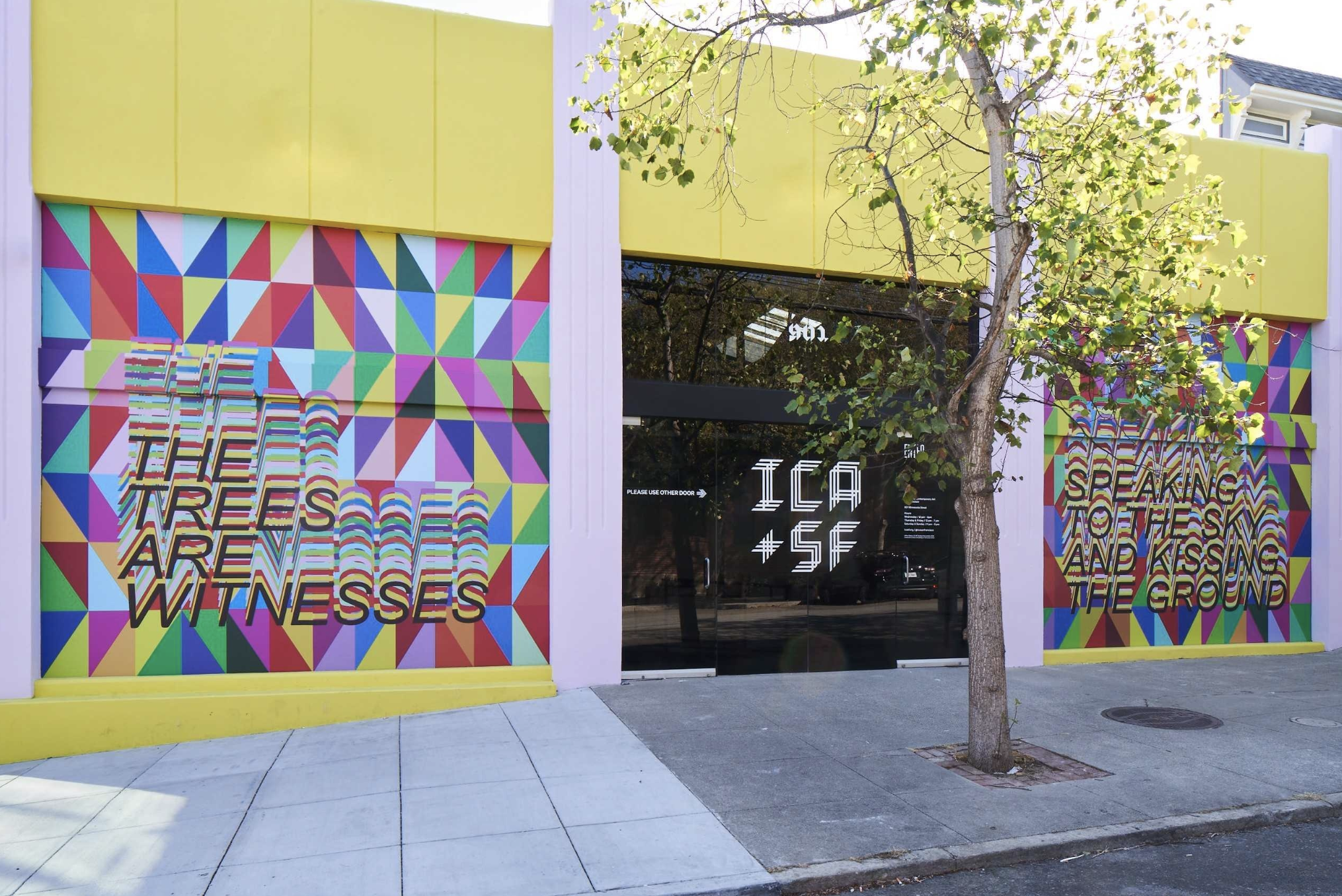 A tree stands in front of the ICA SF building, which is decorated with multicolored, triangular patterns and text that reads "The Trees are Witnesses" and "Speaking to the Sky and Kissing the Ground." Yellow panels are laid horizontally across the top and the bottom of the building.