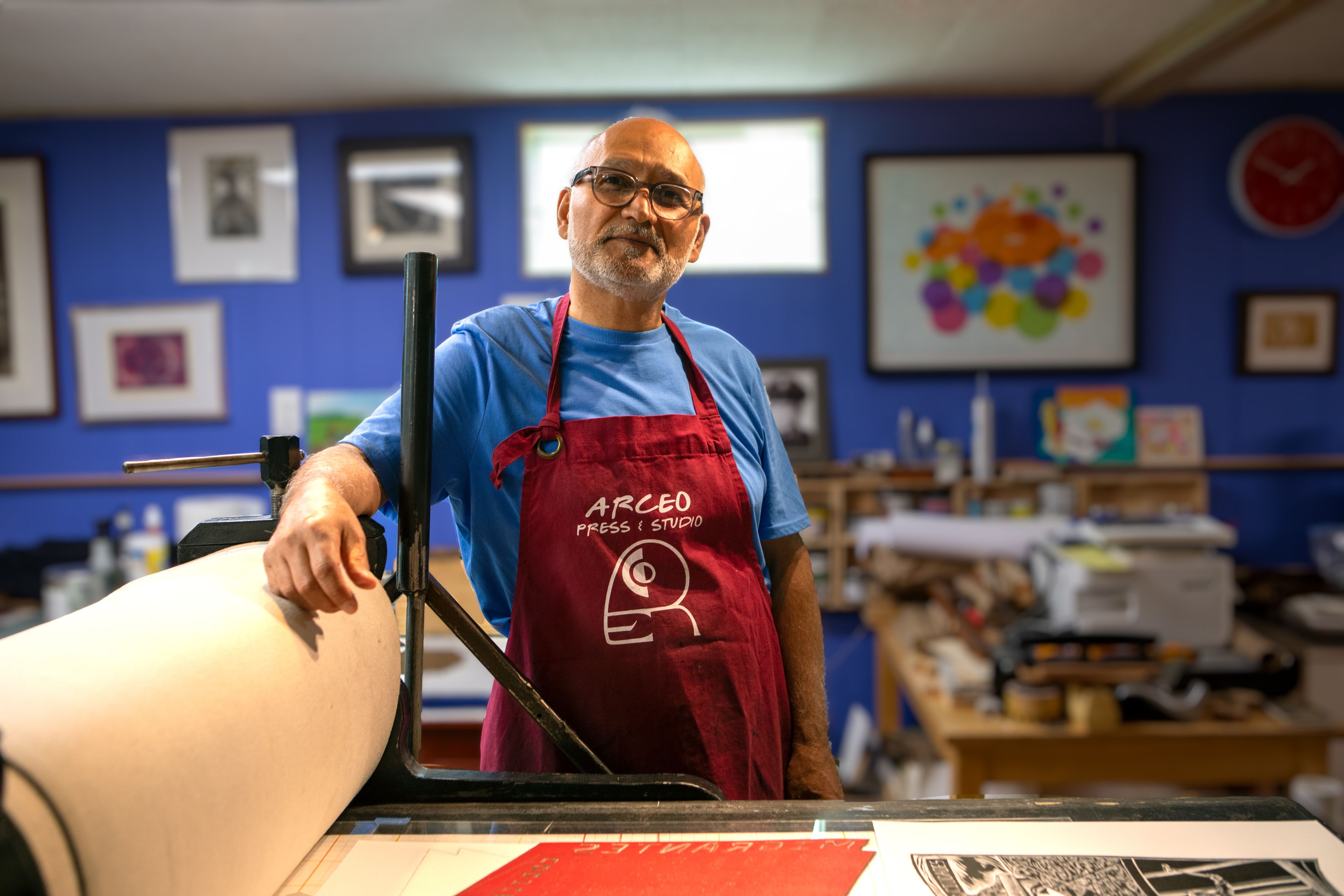 An artist in a red apron stands in front of a press
