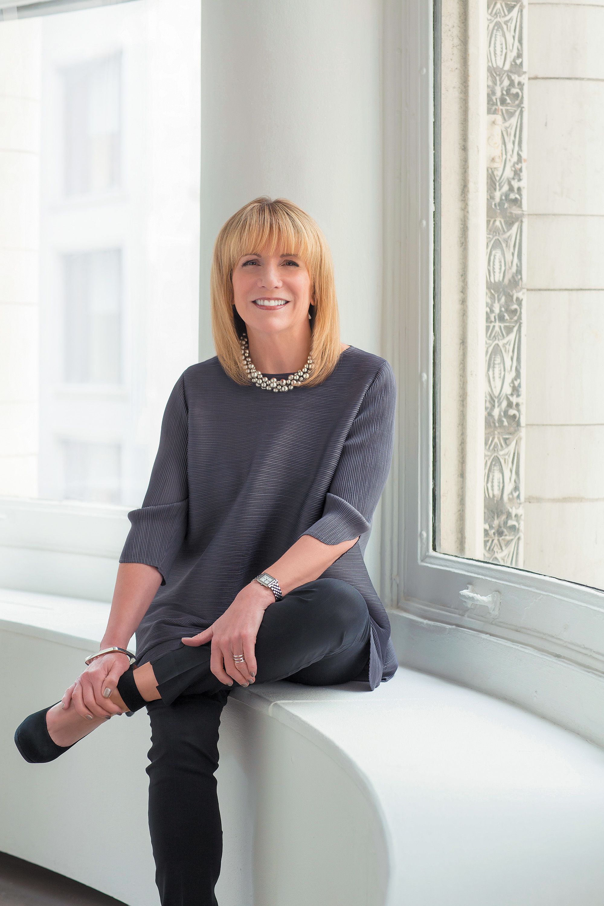 A white woman with shoulder-length blonde hair and straight-across bangs, wearing a chic olive green structured blazer, crosses her arms and smiles into the camera