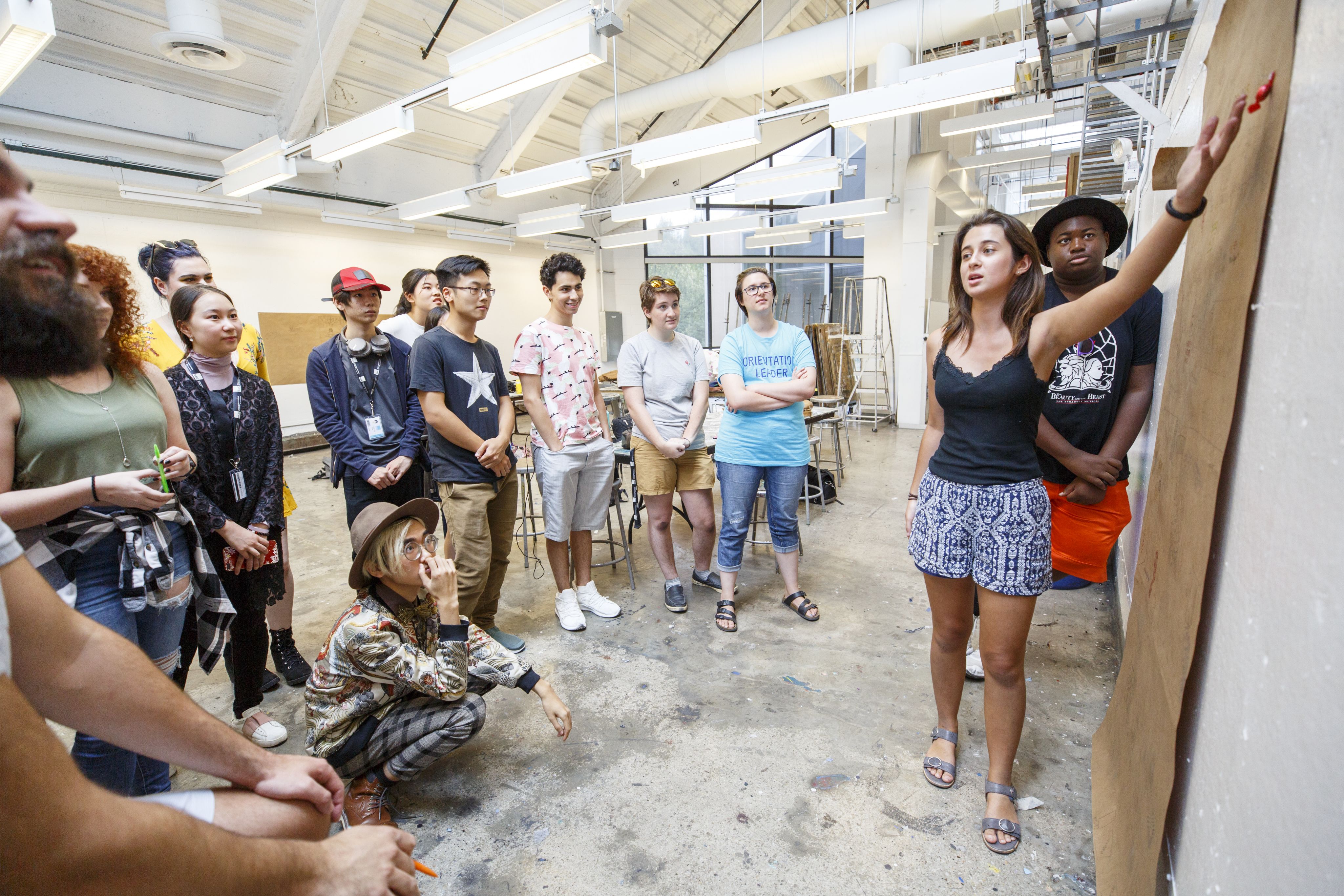 A student gestures at a piece of construction paper on the wall, a group of students surrounding them and listening to her presentation