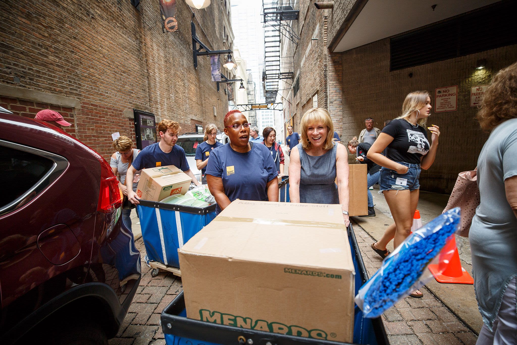 Two women push a cart with a large cardboard box in it down an alley