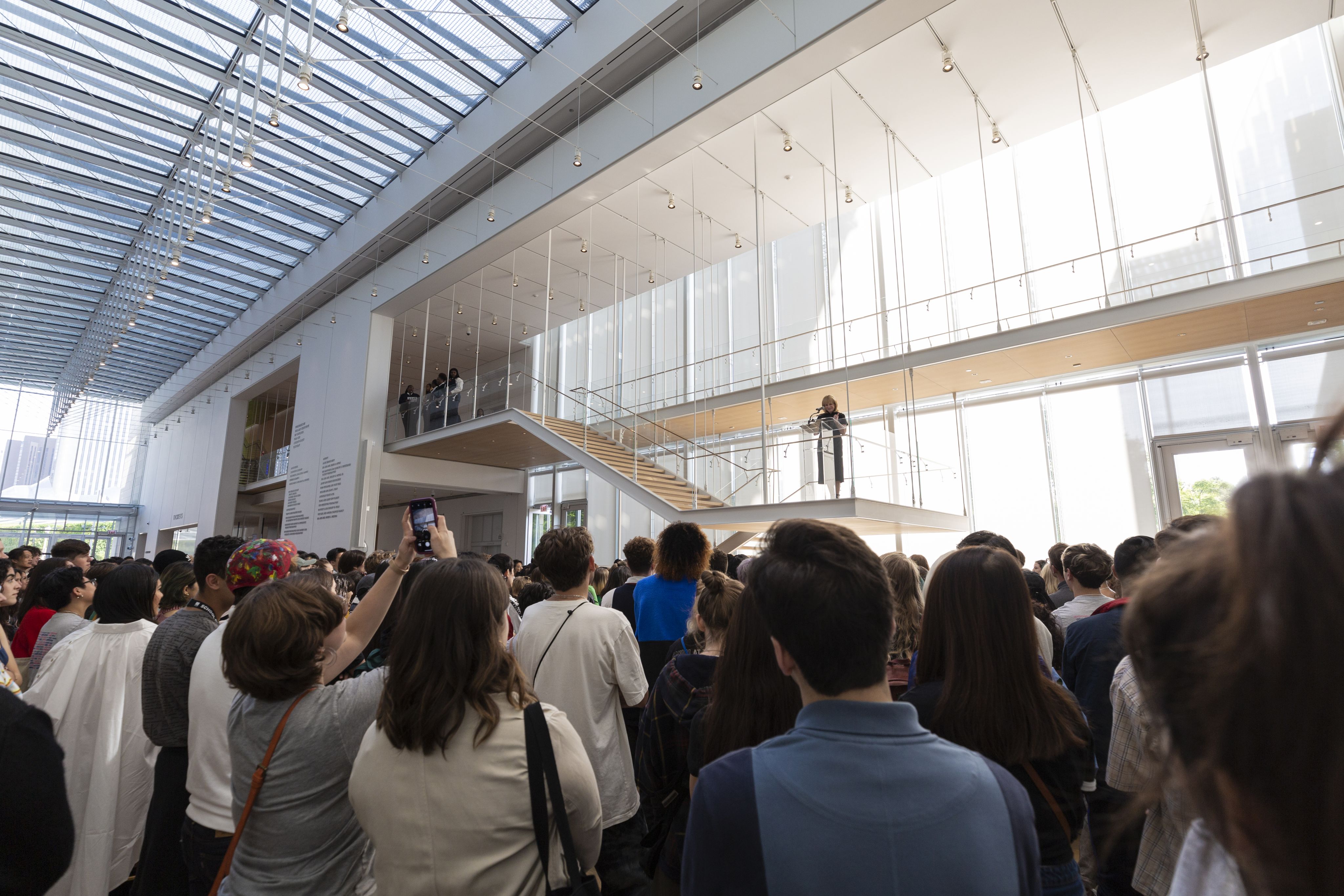 A large crowd gathers in the airy atrium of a museum, looking up at a speaker on a staircase