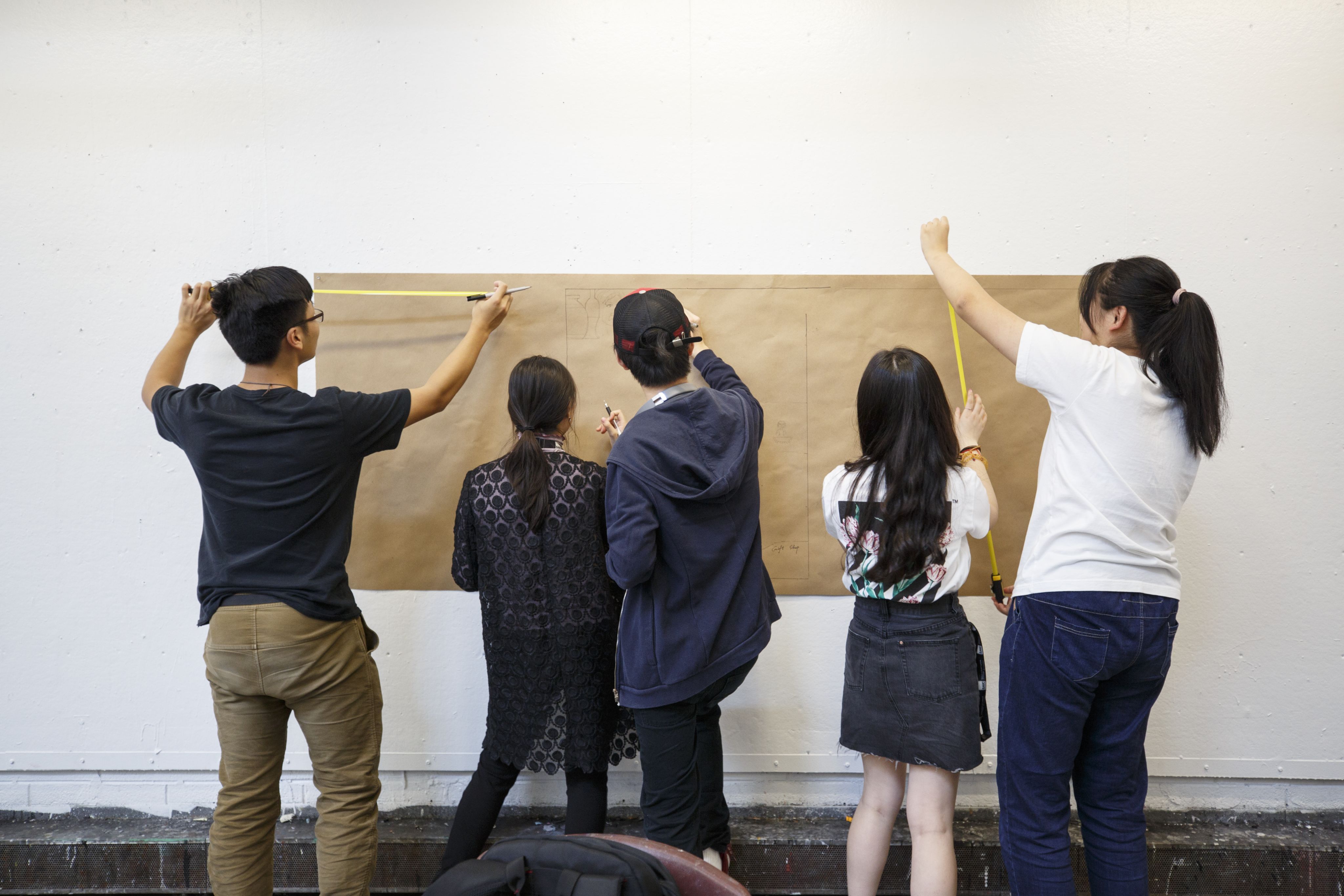 Five students stand in front of a piece of construction paper, two taking measurements