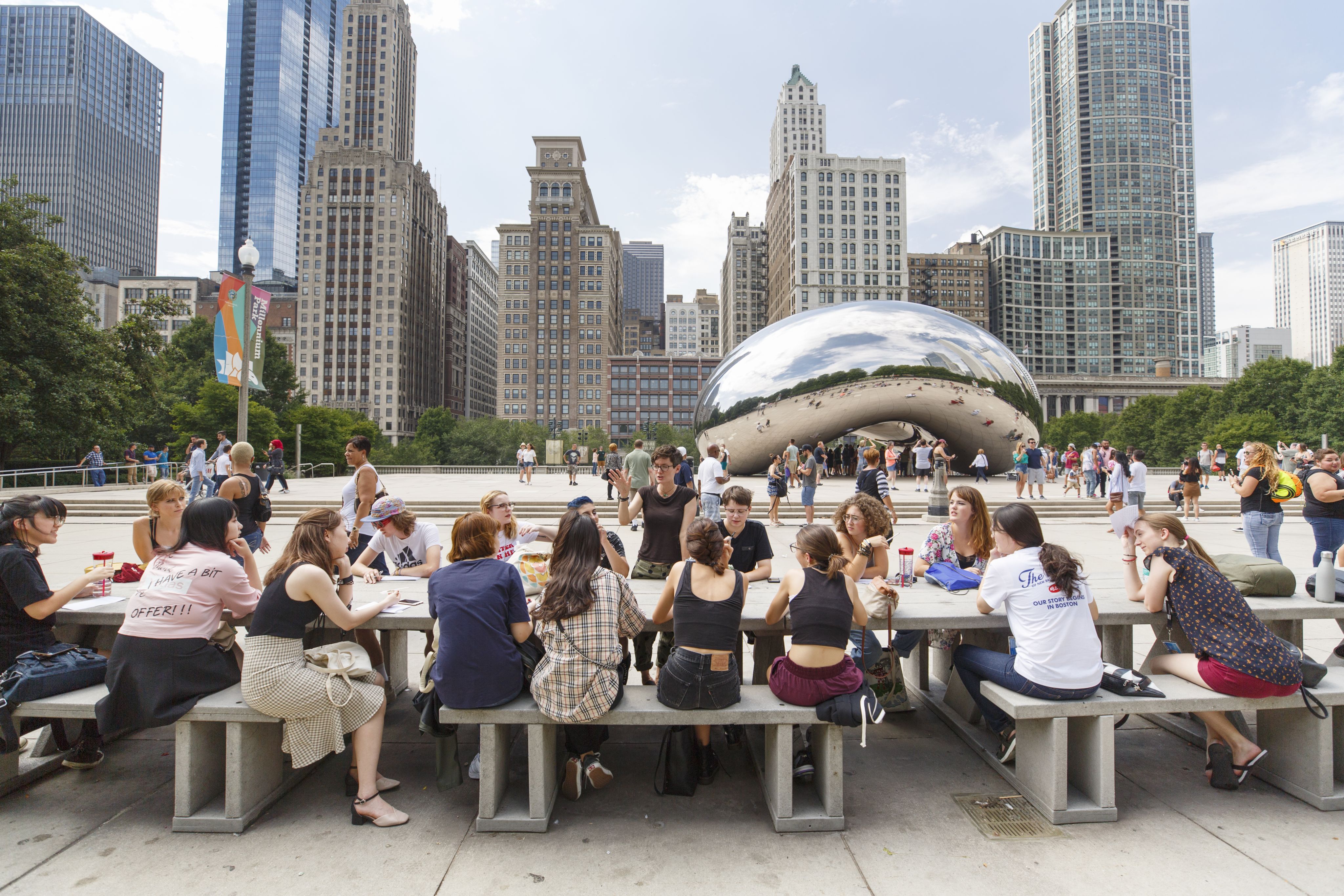 A group of young people sitting at a long table in front of Chicago's Bean sculpture and skyline