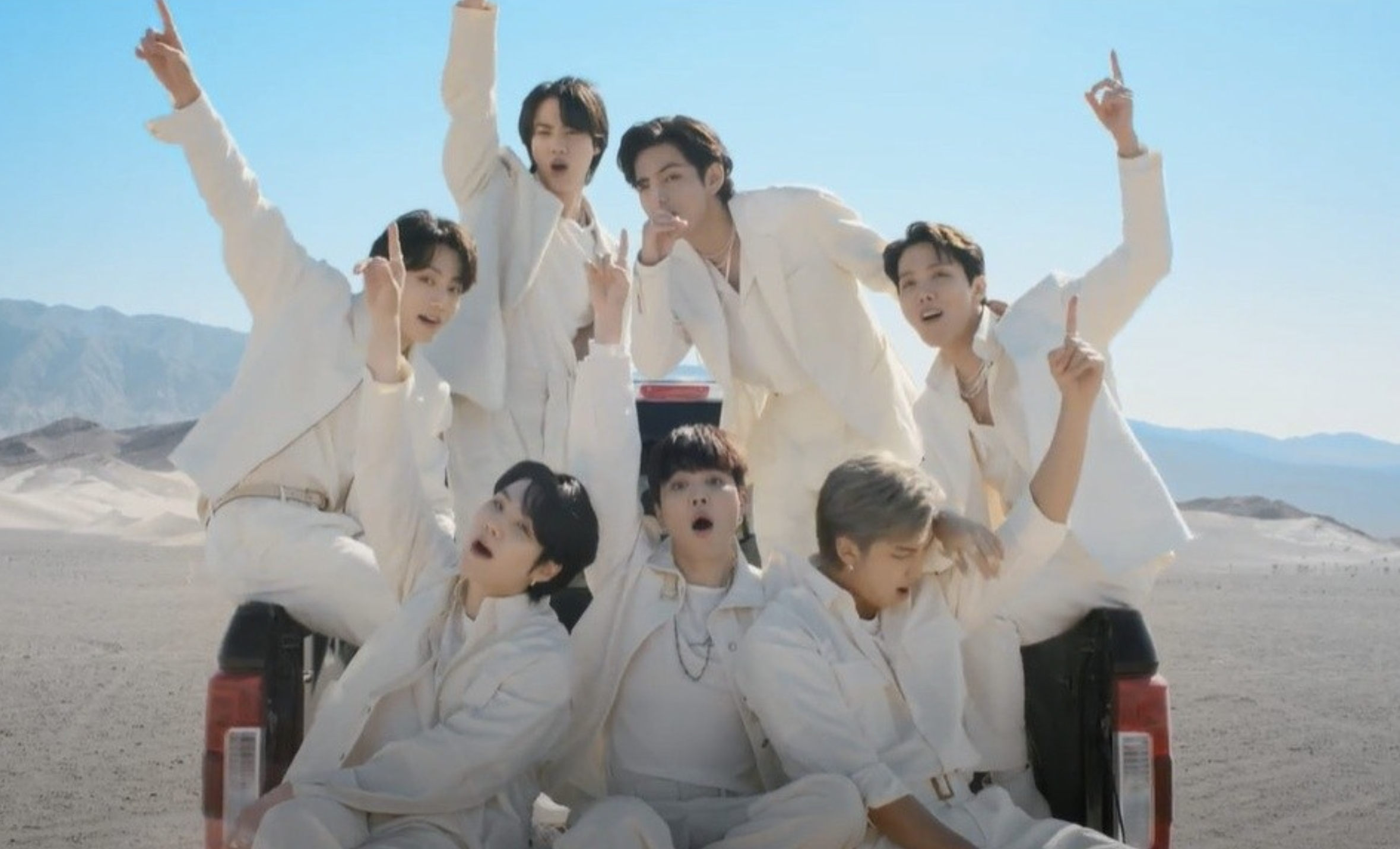 The seven members of BTS sit in the back of a pickup truck. Most of them point their pointer fingers to the sky and look at the camera as they sing. They are all wearing white, and they are located in a white-sand desert with a clear blue sky.