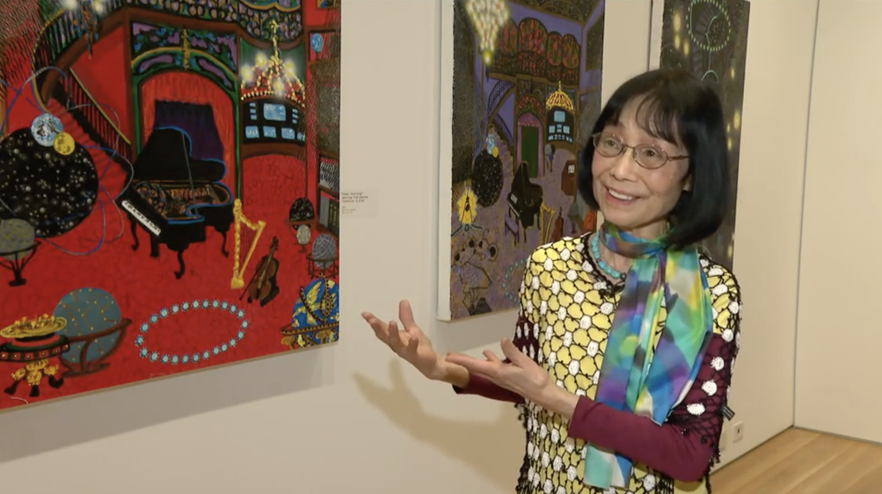 A woman with short black hair stands in front of three paintings and looks to a person off-screen with a smile on her face. She is wearing round glasses, a yellow, green, and purple scarf, and a yellow and magenta shirt with circular patterns.