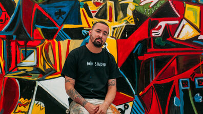An artist stands in front of a colorful work of art in his studio