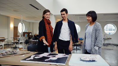 Three people in a large studio room looking at some canvases on a table. 