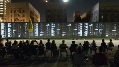 A crowd sitting across the street from a a video projected on a concrete wall at night.