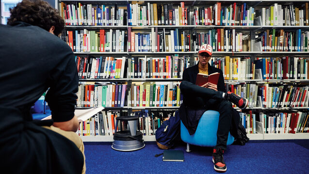  A person reading in a bright blue chair with a large bookshelf behind them. 