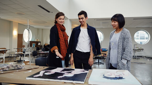 Three people in a large studio room looking at some canvases on a table. 