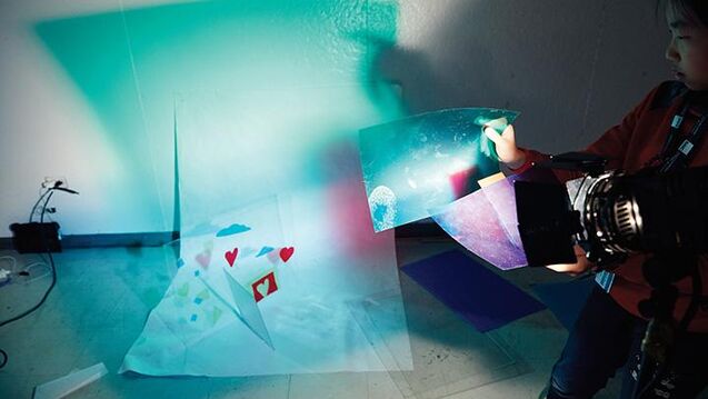 A student using a light with blue tissue paper on the camera lens to create colorful shadow artwork.