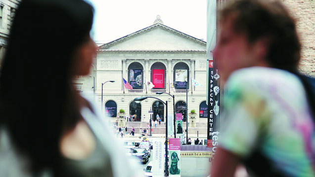 Image of two students in the foreground with the Art Institute of Chicago seen through the windows behind them.