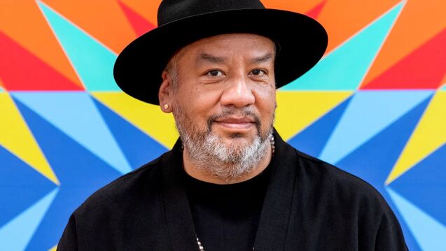 An artist in a black hat poses in front of a colorful, patterned background
