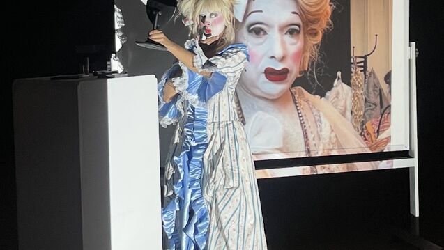 A woman in an elaborate costume stands in front of a screen, emphasizing her unique outfit and the display behind her.