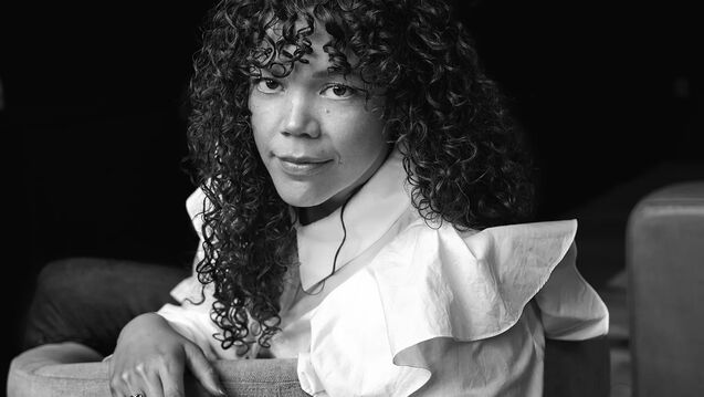 A person with curly hair sits on a chair, wearing a white ruffled shirt, looking towards the camera. The image is in black and white.