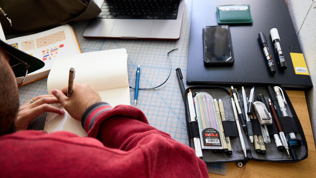 A person's writing by hand in a notebook with pens sitting on the desk.