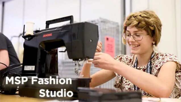 A person with a fair skin tone, glasses and brown hair smiling and sitting at a sewing machine. 
