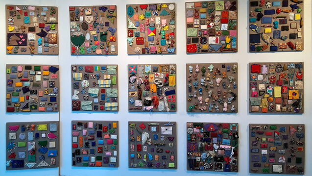 Fifteen square panels mounted on a white wall, each covered with small colorful fabric and beaded bundles arranged in grid-like patterns. Part of Sonya Clark’s The Beaded Prayers Project (1998–present).