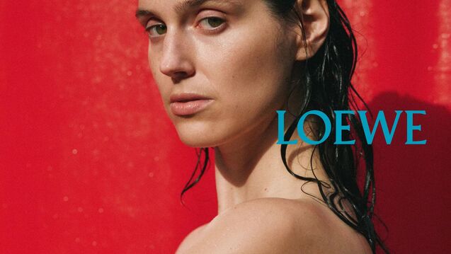 Close-up portrait of actor Eva Victor shown from the shoulders up against a vivid red backdrop, with wet hair and bare shoulders, looking directly at the camera. The word “LOEWE” appears in blue type on the right side of the image.