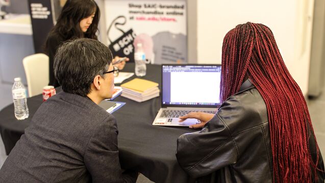 Three people seated at a table, viewing a laptop and a cell phone. 