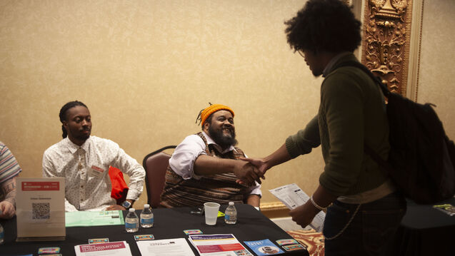 Two adults seated at a table; one of them shaking the hand of another adult standing on the other side. 