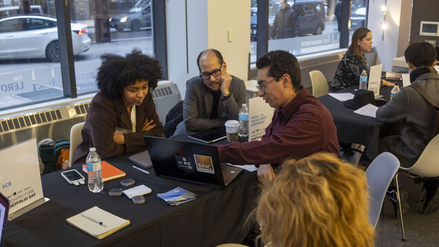 Adults of varying ethnicities and genders gather at tables and view laptops. 